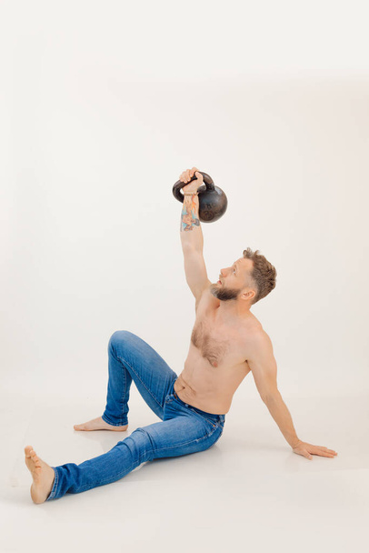 Young athletic man with naked torso sitting and lifting up kettlebell, white background. Keeping fit by strength workout and weightlifting, pumping up muscles. Sport and healthy lifestyle concept. - Foto, Imagen