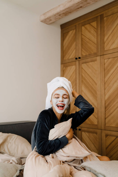 Young woman with face mask laughing while sitting in bed at home - Foto, afbeelding