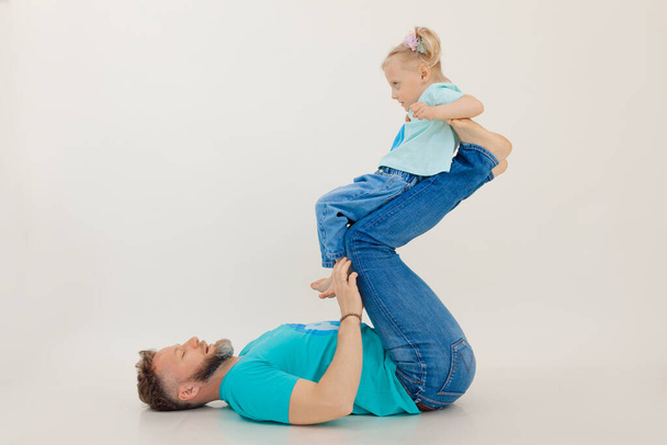 Young athletic man with pumped up arms lying on back holding and lifting up with legs little girl, white background. Whole family workout, father and daughter. Sport and healthy lifestyle concept. - Foto, Bild