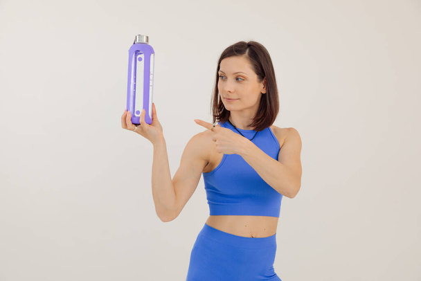 Молодая спортсменка в синем спортивном костюме держит и указывает на бутылку воды, белый фон. Поддержание физической формы в тренажерном зале, накачивание тела. Спорт и здоровый образ жизни. - Фото, изображение