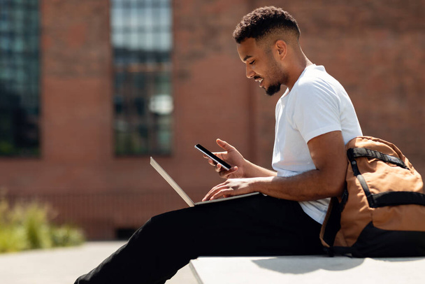 Nuori afrikkalainen amerikkalainen kaveri kannettava tietokone istuu ulkona ja käyttää älypuhelinta, surffata internetissä tai tekstiviestejä, sivunäkymä, kopioi tilaa. Musta mies käyttää modernia kannettavaa tietokonetta ja matkapuhelinta - Valokuva, kuva