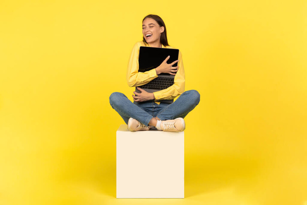 Счастливая леди, обнимающаяся на кубике в студии на желтом фоне. Женщина позирует с компьютером. Фриланс и электронная коммерция, великая реклама - Фото, изображение