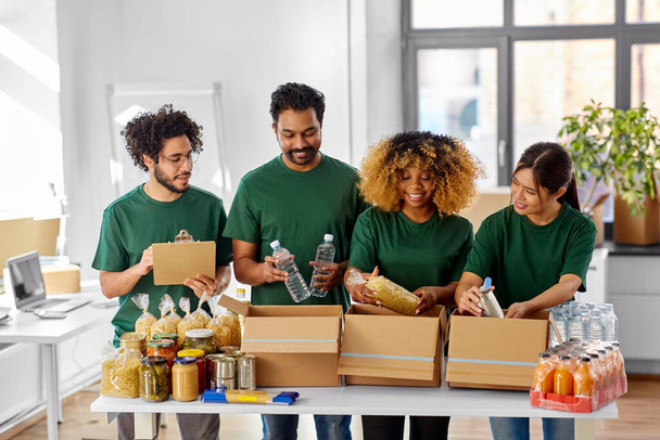 charity, donation and volunteering concept - international group of happy smiling volunteers packing food in boxes according to list on clipboard at distribution or refugee assistance center - Fotoğraf, Görsel