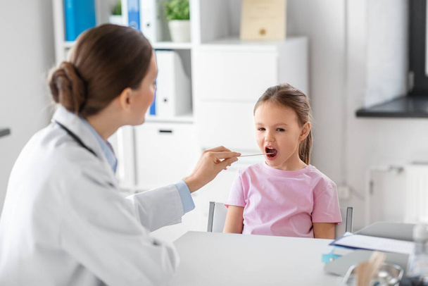 medicine, healthcare and pediatry concept - female doctor or pediatrician with tongue depressor checking little girl patients throat on medical exam at clinic - Foto, immagini