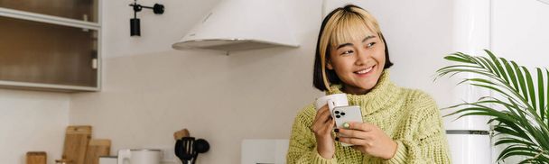 Chica asiática sonriendo y usando el teléfono móvil mientras bebe té en casa - Foto, imagen