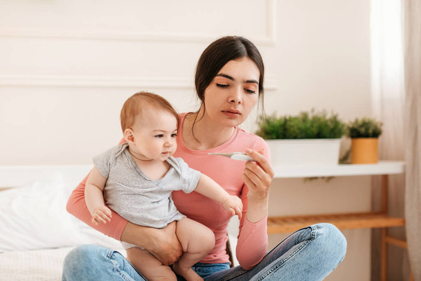 Babies and fever. Young mother measuring kids temperature with thermometer, caring for ill daughter, sitting on bed in bedroom at home. Child care and infants health problem concept - Photo, Image