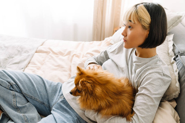 Asian girl petting her ginger dog while resting on bed at home - Photo, Image
