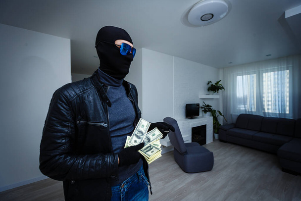 Ένας ληστής ντυμένος με μαύρη κουκούλα στέκεται με μεταμφιεσμένο πρόσωπο και κρατά πολλά χρήματα στα χέρια του, έκλεψε ένα μεγάλο ποσό, ένας κλέφτης έκλεψε μια τηλεόραση. - Φωτογραφία, εικόνα