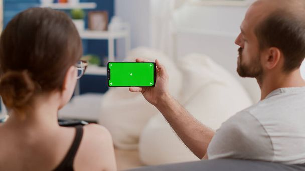 Nahaufnahme eines Mannes, der ein Smartphone mit grünem Bildschirm hält und sich Online-Videoinhalte mit Freundin im modernen Wohnzimmer ansieht. Paar genießt Social-Media-Videos auf Smartphone mit Chroma-Schlüssel. - Foto, Bild