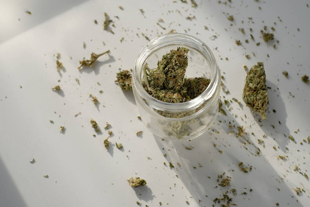 Tienda de marihuana. Tarro de vidrio con cogollos de cannabis secos sobre fondo blanco con marihuana triturada. - Foto, imagen