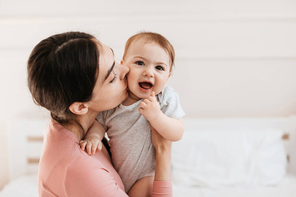 Κοντινό πορτραίτο της αγαπημένης νεαρής μαμάς να φιλάει το μικρό παιδί στο μάγουλο, να δένεται με τον αξιολάτρευτο γιο ή την κόρη της, να κάθεται στο κρεβάτι στο σπίτι, ελεύθερος χώρος - Φωτογραφία, εικόνα