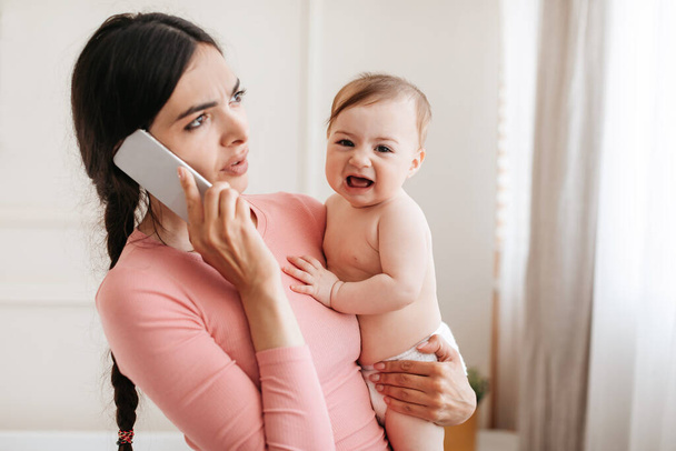 Llama al pediatra. Preocupada joven madre disgustada de llorar bebé, hablando por teléfono celular con el médico y sosteniendo a la pequeña hija en las manos, espacio para copiar - Foto, imagen