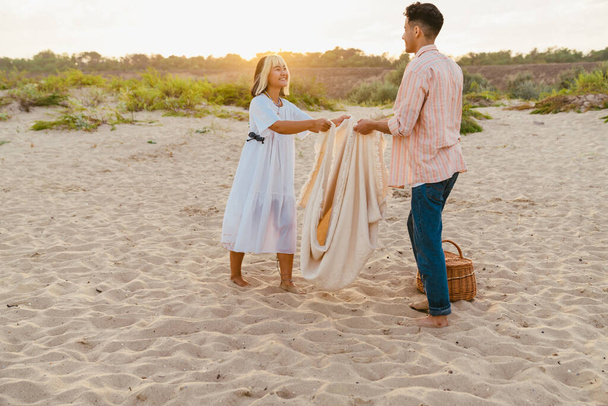 Multiracial couple smiling and holding blanket during picnic on summer beach - Foto, Imagem