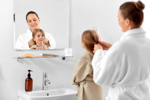 conceito de beleza, família e pessoas - mãe e filha sorridente feliz olhando para espelhar e trançar o cabelo no banheiro - Foto, Imagem