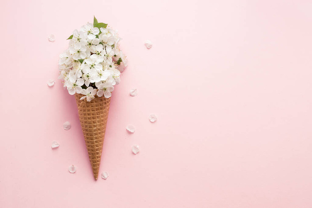 Flat-lay of waffle sweet ice cream cone with white cherry blossom flowers over pastel light pink background. Spring mood minimal concept. Top view. Copy space. - Photo, Image