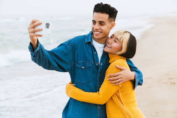 Multiracial young couple taking selfie on cellphone while walking at beach - Fotó, kép