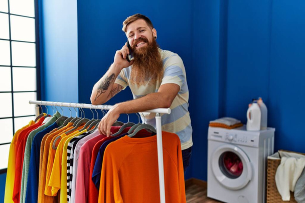 Молодий рудий чоловік посміхається впевнено говорити на смартфоні в пральні
 - Фото, зображення