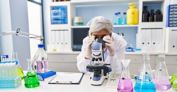 Μεσήλικη γυναίκα που φοράει στολή επιστήμονα χρησιμοποιώντας μικροσκόπιο στο εργαστήριο - Φωτογραφία, εικόνα