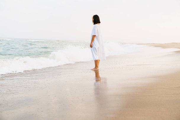 Jovem mulher vestindo vestido olhando para o mar durante a caminhada na praia de verão - Foto, Imagem