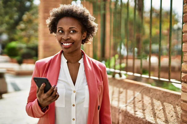 Όμορφη επιχείρηση αφροαμερικανή γυναίκα με αφρο μαλλιά χαμογελαστός χαρούμενος και με αυτοπεποίθηση σε εξωτερικούς χώρους στην πόλη χρησιμοποιώντας smartphone - Φωτογραφία, εικόνα