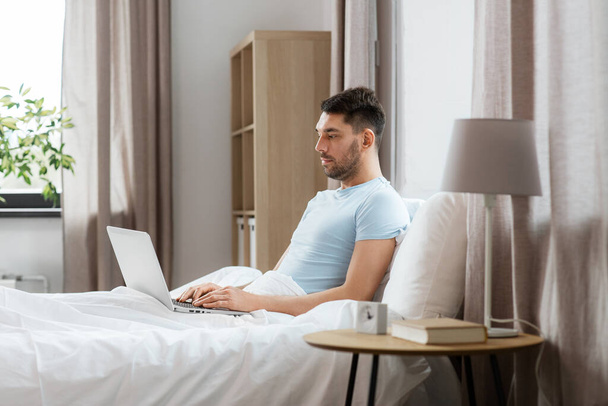 people, technology and remote job concept - man with laptop computer lying in bed at home bedroom - 写真・画像