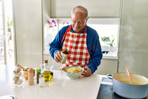 Senior άνθρωπος χαμογελά αυτοπεποίθηση ρίχνει σάλτσα ντομάτας στα μακαρόνια στην κουζίνα - Φωτογραφία, εικόνα