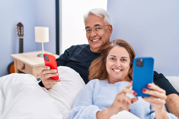 Middle age man and woman couple using smartphone lying on bed at bedroom - Фото, изображение