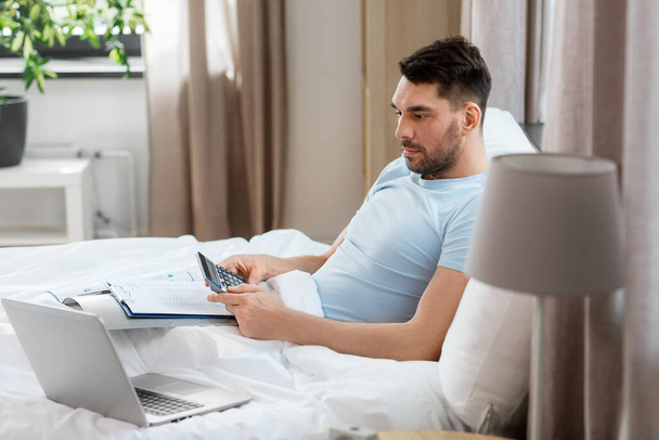 people, technology and remote job concept - man with calculator, papers and laptop computer working in bed at home bedroom - 写真・画像