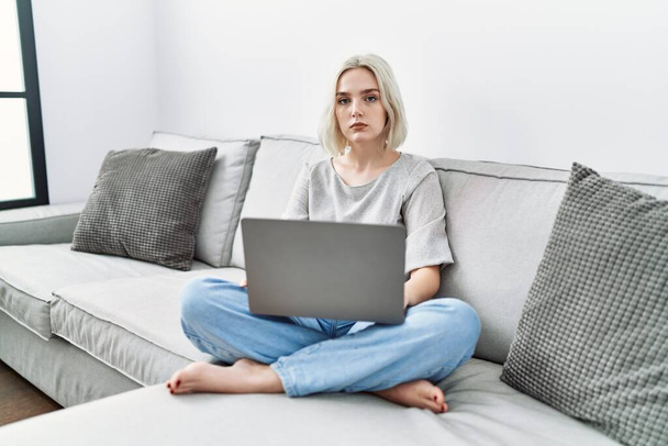 Fiatal kaukázusi nő laptoppal otthon ül a kanapén lazán, komoly arckifejezéssel. egyszerű és természetes nézi a kamerát.  - Fotó, kép