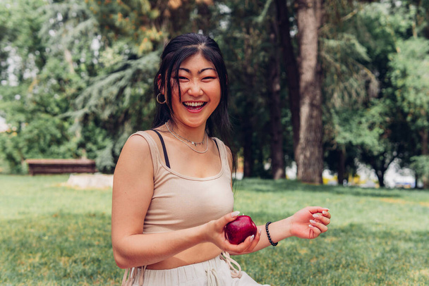 Uśmiechnięta Azjatka z czerwonym jabłkiem na pikniku. Zabawa i zdrowe jedzenie w przyrodzie.Młoda Japonka patrzy w kamerę i śmieje się z pikniku z przyjaciółmi jedząc owoce. - Zdjęcie, obraz