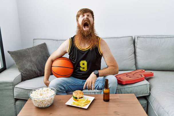 Caucasian man with long beard holding basketball ball cheering tv game angry and mad screaming frustrated and furious, shouting with anger. rage and aggressive concept.  - Foto, afbeelding