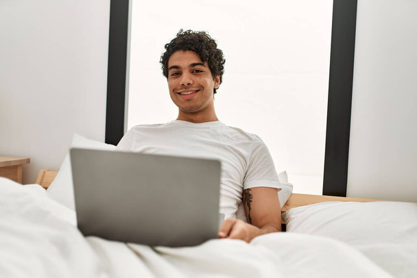 Mladý hispánec pomocí notebooku sedí na posteli v ložnici. - Fotografie, Obrázek