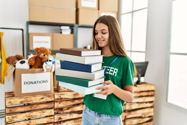 Adorable girl wearing volunteer uniform holding books at charity center - Photo, image