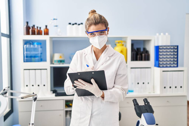 Νεαρή ξανθιά γυναίκα φορώντας στολή επιστήμονα και ιατρική μάσκα γραφής στο πρόχειρο στο εργαστήριο - Φωτογραφία, εικόνα