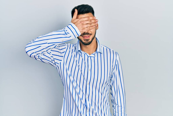 Young hispanic man with beard wearing casual striped shirt smiling and laughing with hand on face covering eyes for surprise. blind concept.  - Photo, Image