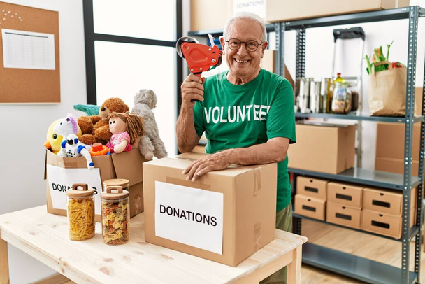 Ανώτερος άνδρας φορώντας στολή εθελοντή συσκευάζοντας χαρτόκουτα δωρεών σε φιλανθρωπικό κέντρο - Φωτογραφία, εικόνα