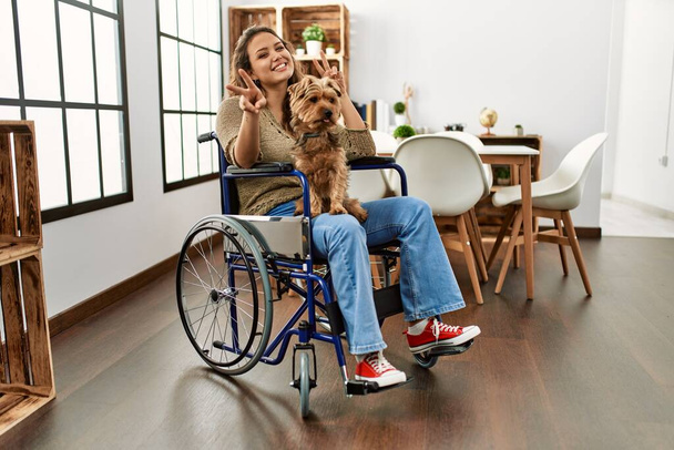 Menina hispânica jovem sentado em cadeira de rodas em casa sorrindo positivo fazendo sinal ok com a mão e os dedos. expressão bem sucedida.  - Foto, Imagem