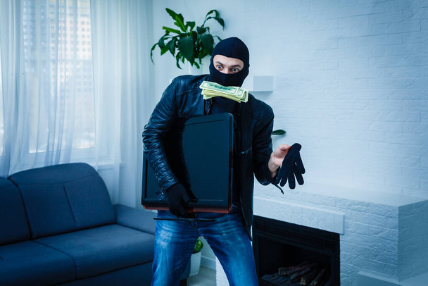 Robber man dressed in black hoodie stands with disguised face and holds a lot of money in his hands, stole a large amount, a thief man stole a TV. - Foto, Bild