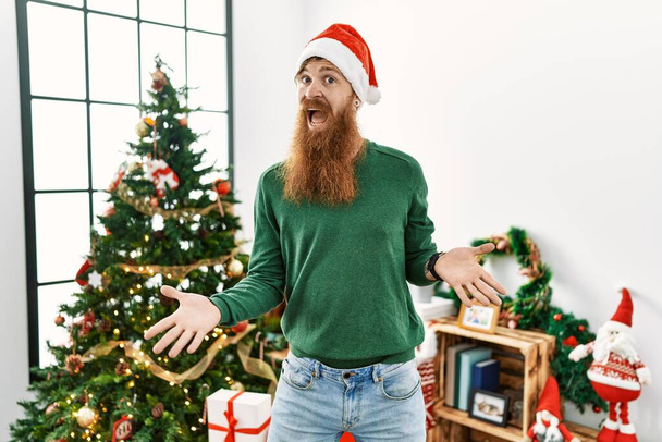 Рыженький с длинной бородой, в рождественской шляпе, рядом с рождественской ёлкой, веселый, с распростертыми объятиями, приветливый, позитивный и уверенный в себе привет  - Фото, изображение