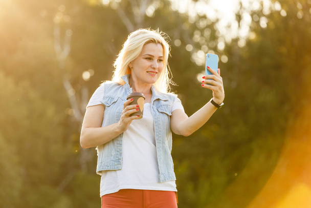 Beautiful happy young woman taking a selfie photo with smart phone outdoors in park on summer day. Modern millennial youth lifestyle and travel concept - Zdjęcie, obraz