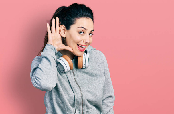 Νεαρή ισπανίδα γυναίκα που φοράει ρούχα γυμναστικής και χρησιμοποιεί ακουστικά χαμογελώντας με το χέρι πάνω από το αυτί ακούει και ακούει φήμες ή κουτσομπολιά. έννοια της κώφωσης.  - Φωτογραφία, εικόνα