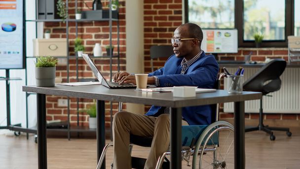 Gelähmter Mitarbeiter im Rollstuhl mit Laptop zur Planung von Start-up-Projekten. In einem behindertengerechten Büro leiden sie unter chronischer Beeinträchtigung und körperlichem Zustand. Stativschuss. - Foto, Bild
