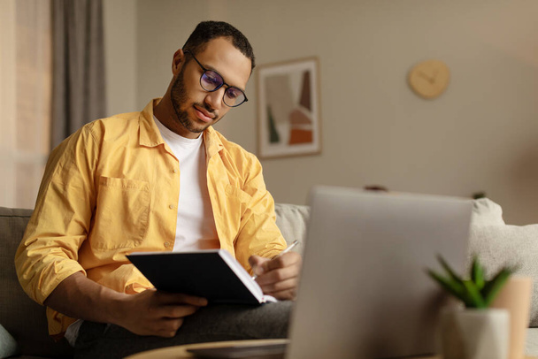 Focused young African American man using laptop, working online, taking notes during business meeting at home, free space. Millennial black guy learning online, writing down info - Foto, afbeelding
