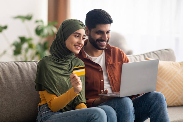 Boldog közel-keleti házastársak Online vásárlás Laptop Számítógépen keresztül Bankkártyával történő fizetés Internet Banking szolgáltatások használatával Együtt ülnek a kanapén otthon. Oldalnézet - Fotó, kép