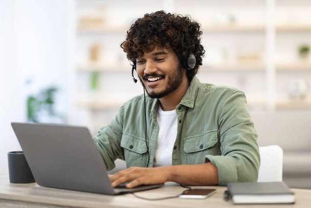 Positive handsome young indian guy independent contractor working online from home, sitting at workdesk, using laptop and headset, having video call with business partner, copy space - Foto, immagini