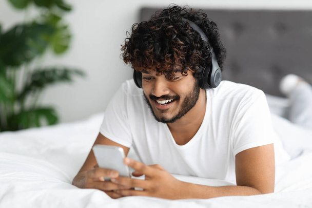 Boldog vonzó, fiatal, sötét bőrű srác modern okostelefont és vezeték nélküli fejhallgatót használ, miközben otthon lehűl az ágyban, videókat néz az interneten, videojátékot játszik, másolja a helyet, közelről - Fotó, kép