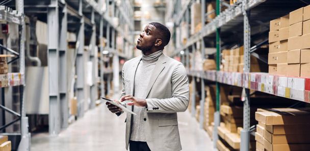 Portret van lachende Afrikaans-Amerikaanse zakenman bestel details op tablet controleren van goederen en benodigdheden op rekken met goederen achtergrond in warehouse.logistic en zakelijke export - Foto, afbeelding
