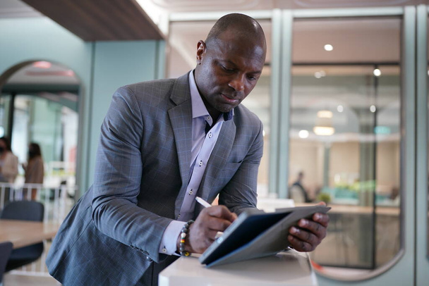 Portret Afroamerykańskiego biznesmena korzystającego z laptopa urządzenia mobilnego w nowoczesnym biurze. - Zdjęcie, obraz