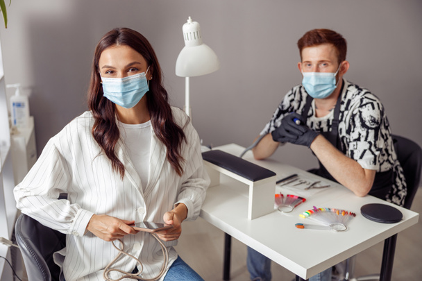 Vrolijke vrouw client dragen beschermende masker en nagel technicus kijken naar de camera terwijl zitten op het werk tafel in nagel studio - Foto, afbeelding