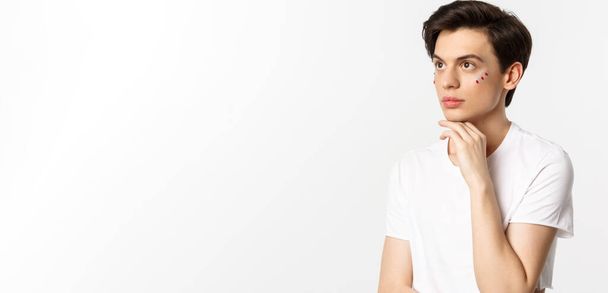 People, lgbtq and beauty concept. Beautiful androgynous man with glitter under eyes, looking at upper left corner, posing against white background. - Foto, Bild